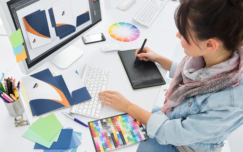Graficzka projektuje teczkę firmową na tablecie przy monitorze i próbkach kolorów
