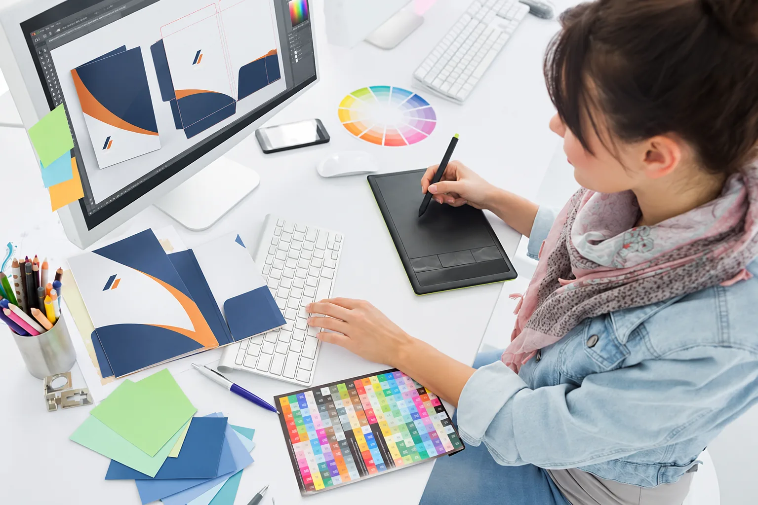 Graficzka projektuje firmową teczkę przy monitorze, tablecie i próbkach kolorów